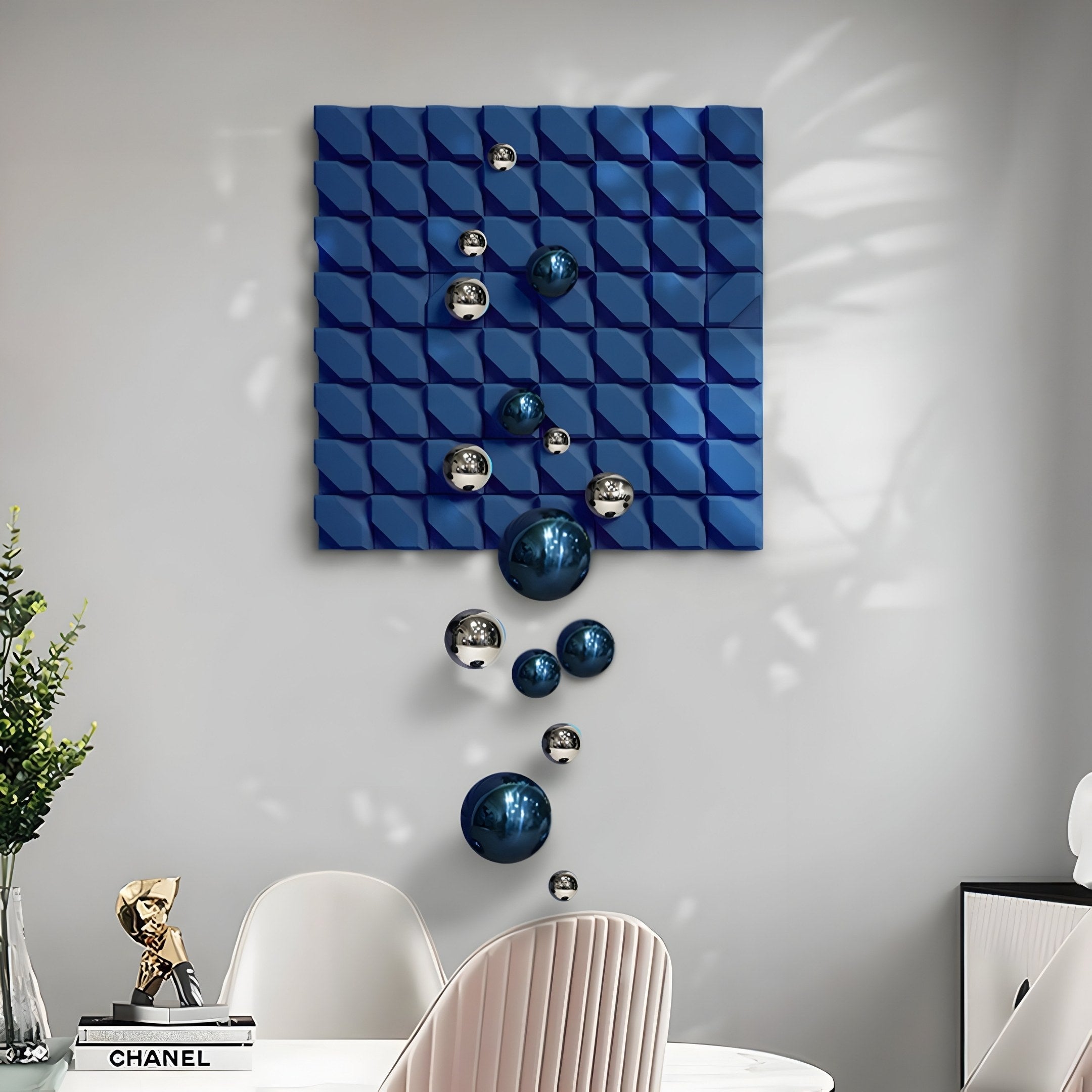 A modern office features a white table with minimalist decor, including a plant and Chanel book. Complementing the space, the Orbina Blue 3D Wood Relief Wall Art by Giant Sculptures showcases textured wood with silver and blue reflective spheres in various sizes.