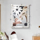 The Florere Neutral Bloom Hand-Painted Sandstone 3D Wall Art by Giant Sculptures features abstract floral designs in white, beige, and brown tones with large daisy-like flowers and geometric shapes. It is elegantly displayed above a leather chair next to a wooden side table decorated with a plant.