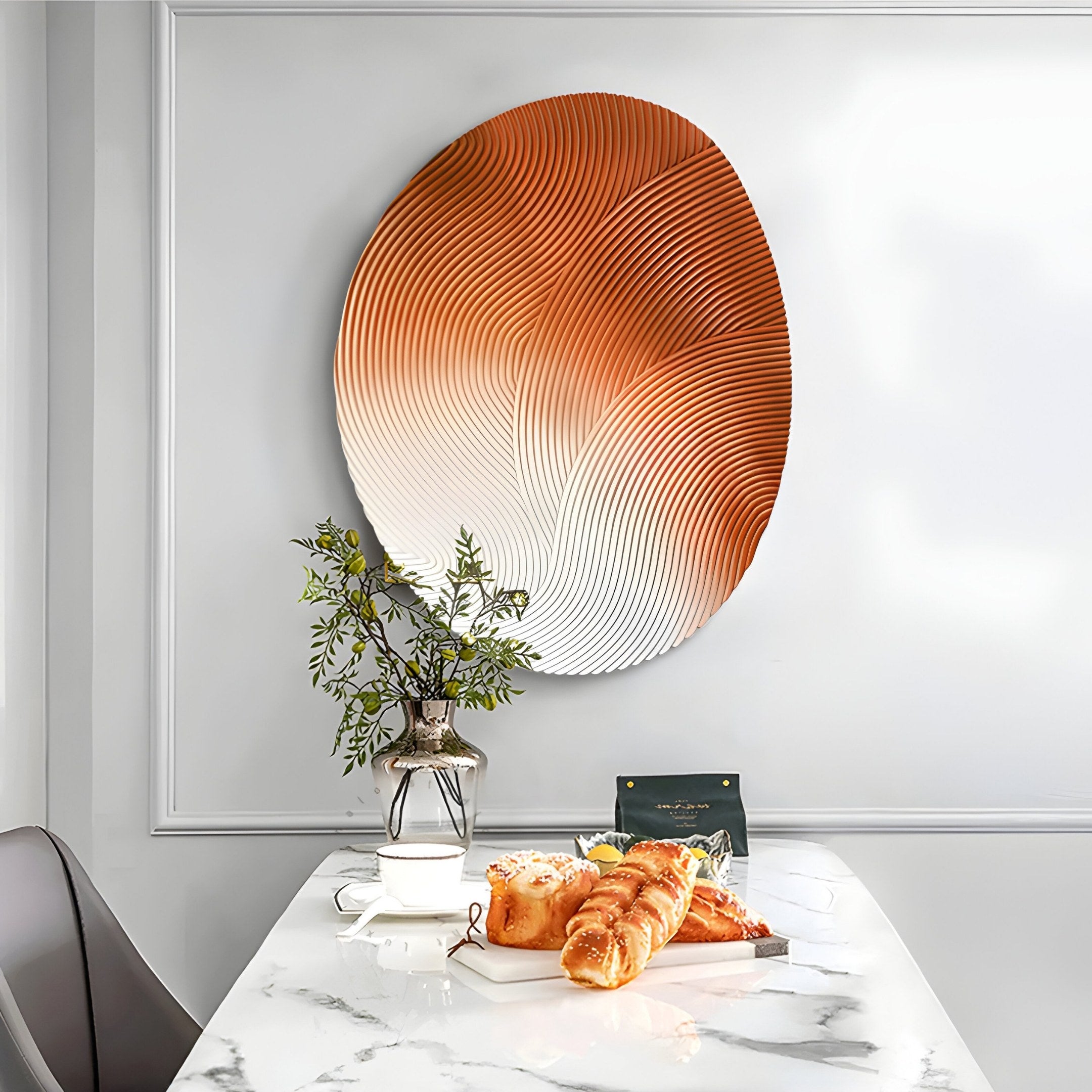 In a modern dining area, a marble table set with croissants and a small cake is complemented by a vase-held plant. The wall displays Giant Sculptures Fluctus Orange Textured Sandstone Wood Relief 3D Wall Art, showcasing abstract orange and white patterns for added depth and texture.