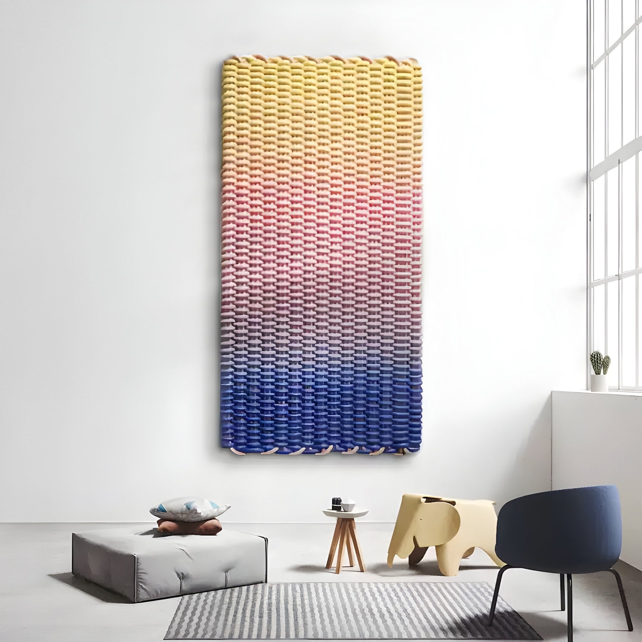 A modern room showcases the Spectrum Weave Vertical Sunset Hues Wood Relief 3D Wall Art by Giant Sculptures, with a gray cushion and plush toys, a small wooden table holding a book, and a blue chair near a tall window.