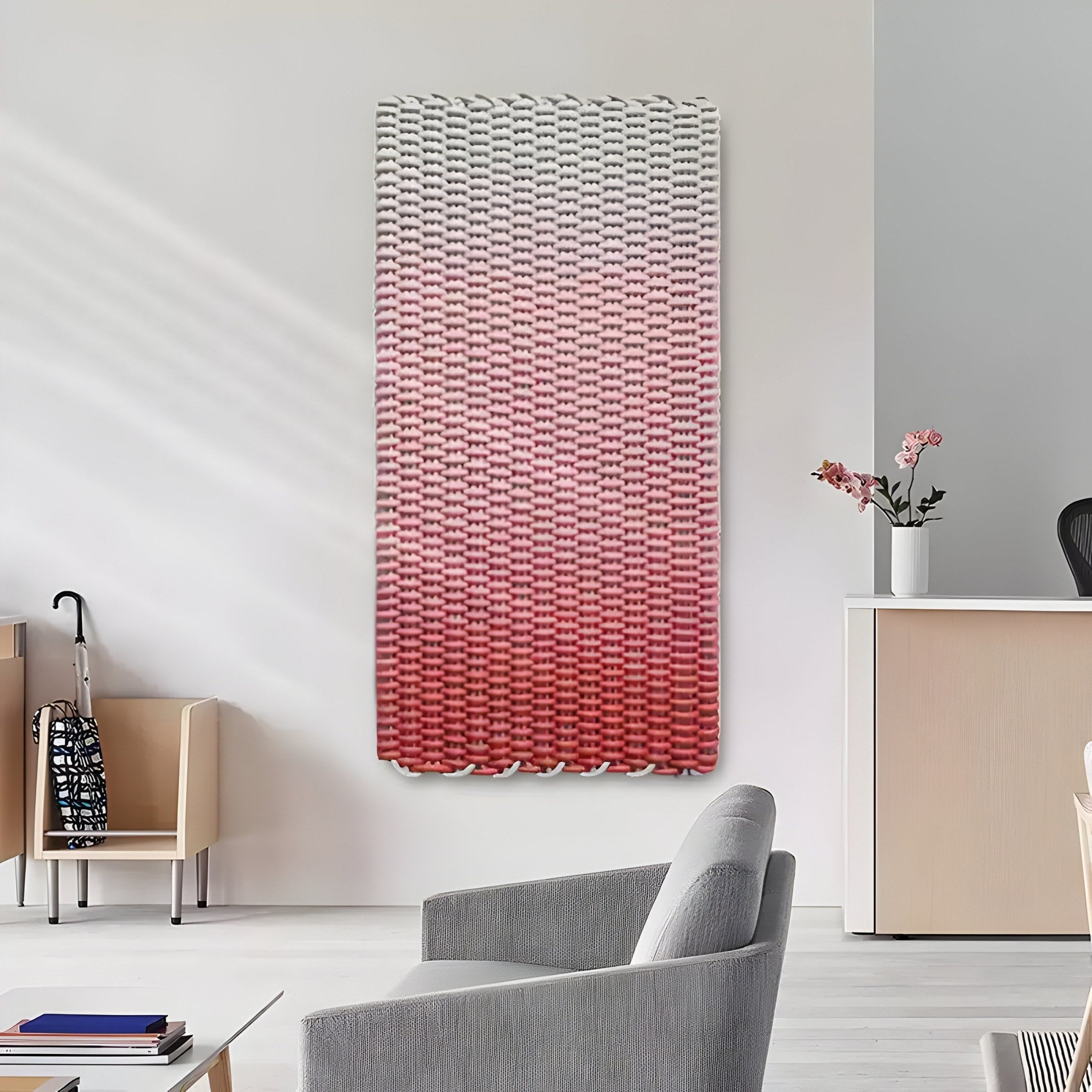 A modern office lobby with light-colored walls and furniture features the stunning Spectrum Weave Vertical Rose Blush Wood Relief 3D Wall Art by Giant Sculptures. A small table with pink flowers completes the aesthetic, while a backpack rests casually on a chair in the corner.