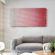 A modern living room showcases a gray textured sofa against a light wall, highlighted by Spectrum Weave Horizontal Rose Blush Wood Relief 3D Wall Art by Giant Sculptures. Abstract woven art in red and white hues and a small wooden table with black metal base add texture, complemented by white decor pieces for coziness.