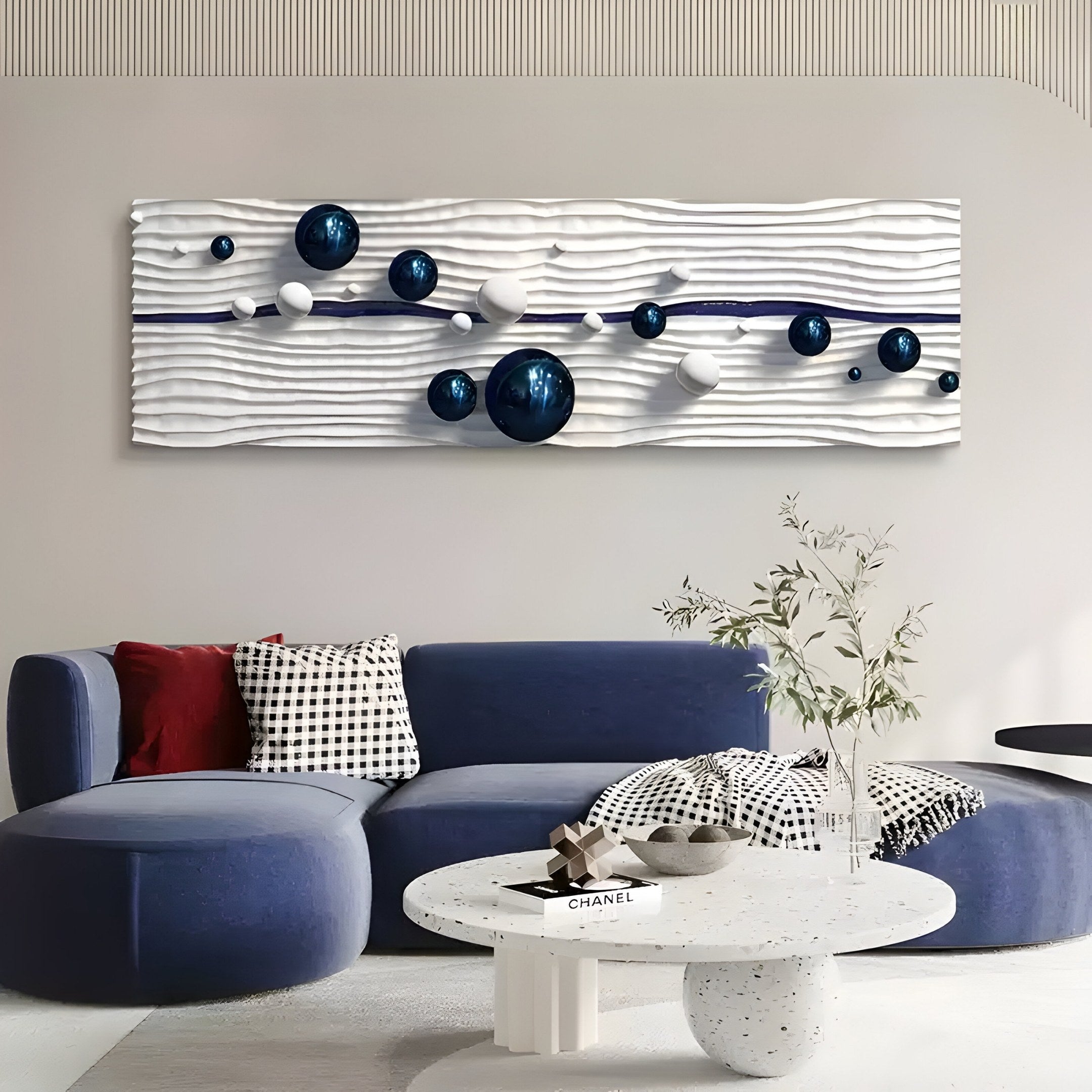 A modern living room features a curved blue sofa with red and black-and-white pillows. Above, Giant Sculptures Lustra Sapphire Blue & White 3D Wood Relief Wall Art With Metal Ball showcases spheres on a textured white background. A round white terrazzo coffee table with a vase and book completes the elegant space.