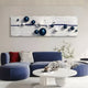 A modern living room features a curved blue sofa with red and black-and-white pillows. Above, Giant Sculptures Lustra Sapphire Blue & White 3D Wood Relief Wall Art With Metal Ball showcases spheres on a textured white background. A round white terrazzo coffee table with a vase and book completes the elegant space.