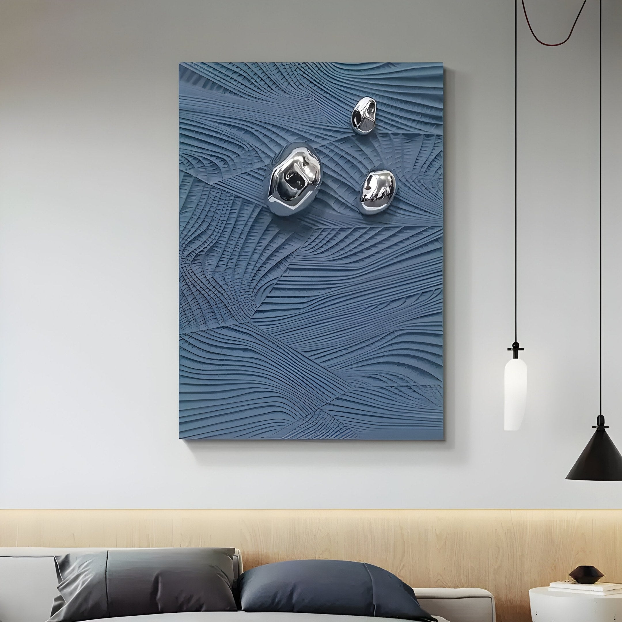A contemporary living room features the Verdara Blue Wood Relief 3D Wall Art by Giant Sculptures, exhibiting textured patterns and metallic accents. A light wooden ledge below displays dark and light pillows. Overhead, a white and a black pendant light illuminate the space.