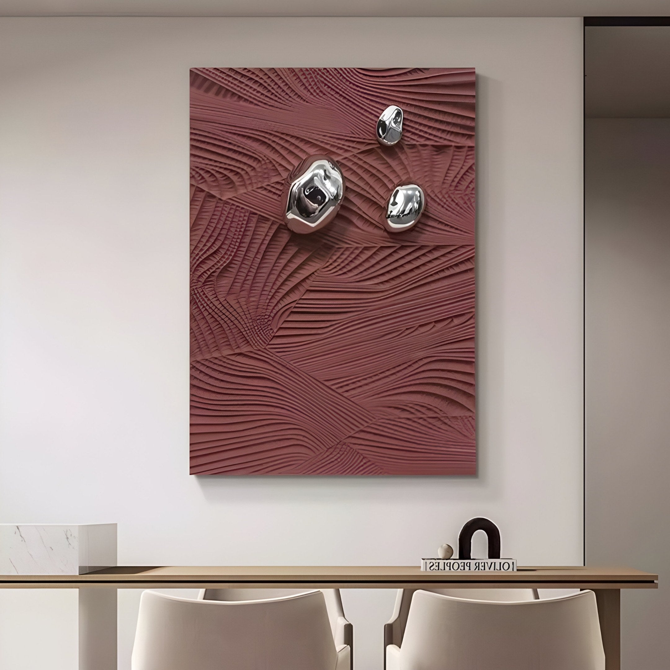 The Verdara Burgundy Red Wood Relief 3D Wall Art by Giant Sculptures boasts a textured surface with metallic spheres. Hanging above a light wood bench and two white chairs, it enhances a small minimalistic shelf holding books and a black bowl.