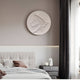 In a modern, minimalist bedroom, light gray bedding with a brown pillow complements the Selene I White Sandstone Textured 3D Wood Relief Wall Art by Giant Sculptures above. A dark curtain partially veils a large window beside a gray nightstand topped with a small sculpture and sphere.