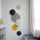 The Orbita 6-Set Circular Neutral Grey Wall Art by Giant Sculptures displays textured panels in black, white, beige, and yellow. With a sheer-curtained window nearby and a blue-cushioned bench plus wire-frame shelf with flowers, it enhances this stylish interior.