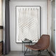 A contemporary room showcases a Giant Sculptures Elorium White Concentric Hand-Painted Wood Relief 3D Wall Art, complementing a modern brown lounge chair and small gray side table with decor. Floor-to-ceiling curtains frame a window on the left, enhancing the minimalist design.