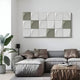 A modern living room features a gray sectional sofa with brown cushions and an ottoman. Nearby, a potted plant complements the Averra White & Sage Block Wood Relief 3D Wall Art by Giant Sculptures, which elegantly decorates the wall above the sofa.