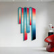 The Giant Sculptures Opaline Coral Teal Gradient Wood Relief 3D Wall Art displays multicolored vertical panels transitioning from red to blue, mounted on a light gray wall, with a modern red ribbed chair on the right and a small sculpture atop a white pedestal.