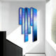 The Opaline Twilight Azure Gradient Wood Relief 3D Wall Art by Giant Sculptures features elongated vertical panels in blue and purple hues, casting shadows on a white wall in natural light. A staircase with a black railing is partially visible.