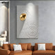 A modern living room features a brown sofa with white cushions. The wall is highlighted by the Ondella Gold Ripple I Sandstone Wood Relief 3D Wall Art by Giant Sculptures, set against concentric circles on a textured gray backdrop. A niche sculpture and soft lighting complete the ambiance.