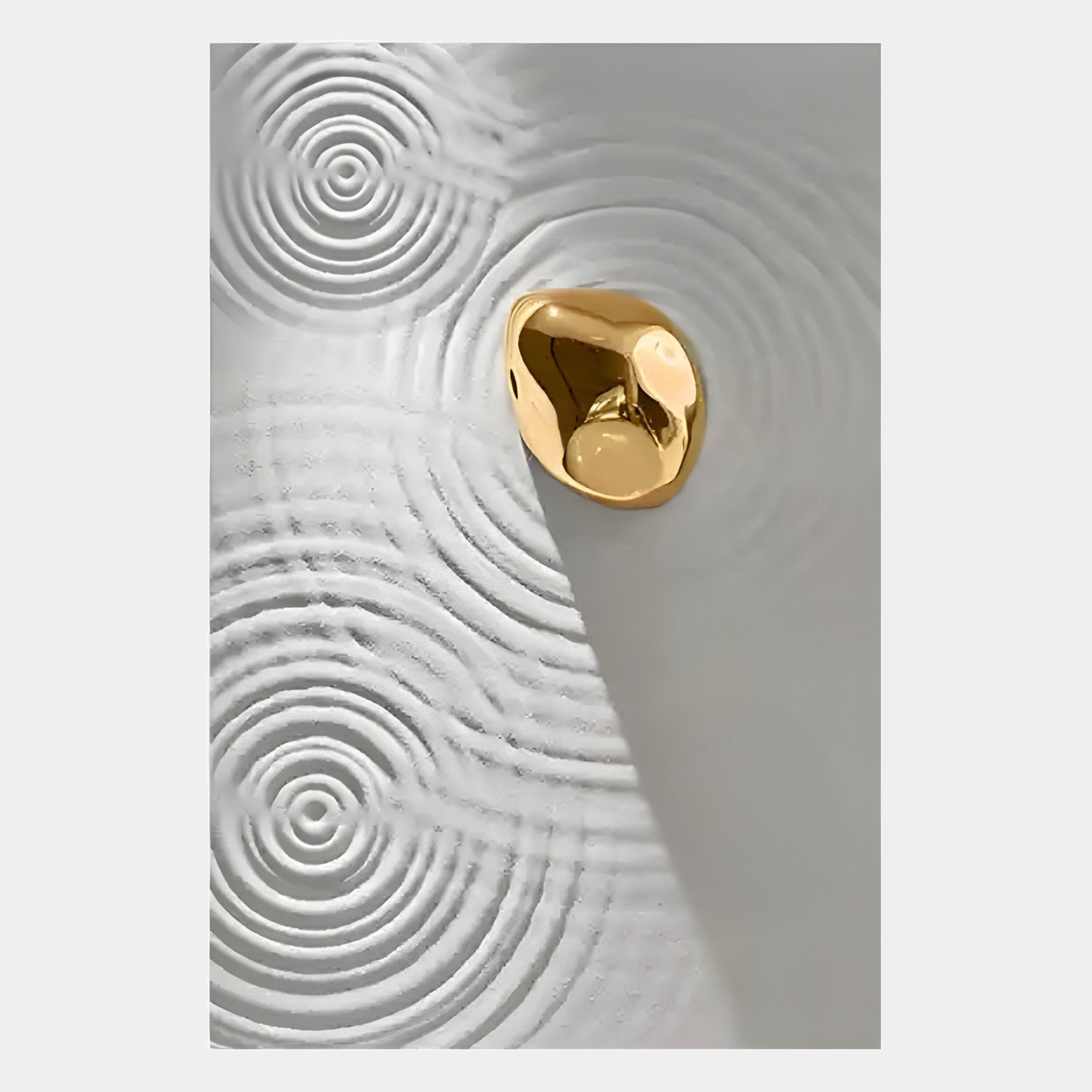 A shiny gold pebble rests on a smooth white surface, casting a shadow amid circular patterns like water ripples—this is the Ondella Gold Ripple II Sandstone Wood Relief 3D Wall Art by Giant Sculptures.