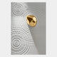 A shiny gold pebble rests on a smooth white surface, casting a shadow amid circular patterns like water ripples—this is the Ondella Gold Ripple II Sandstone Wood Relief 3D Wall Art by Giant Sculptures.