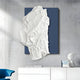 Displayed on a concrete wall is the Veritas White & Blue Abstract Wood Relief 3D Wall Art by Giant Sculptures, resembling crumpled paper. Below is a white cabinet with books and a modern lamp, with a blue chair partially visible in the corner.