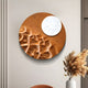The Verve Earthy Brown Wood Relief 3D Wall Art by Giant Sculptures features a round, moon-like design with a smaller white circle. It hangs on a light wall above a ceramic vase of dried plants, with an orange chair partially visible in the corner.