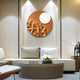 A modern living room features a circular coffee table and light-gray sofas. On the wall, an eye-catching Verve Earthy Brown Wood Relief 3D Wall Art by Giant Sculptures is illuminated by two small lamps. A vase of white flowers on the table highlights the contemporary design.