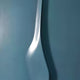 The Velar Vertical Blue Wood Carving 3D Wall Art by Giant Sculptures is a metallic, abstract piece with wavy designs. It reflects light against a smooth teal wall, creating soft highlights and shadows that accentuate its curvature and texture.