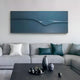 In a modern living room, a light gray sectional sofa with white and teal pillows is complemented by the Velar Horizontal Blue Wood Carving 3D Wall Art from Giant Sculptures showcasing a wave motif above it. A dark coffee table adorned with decorative vases and ornaments completes the scene.