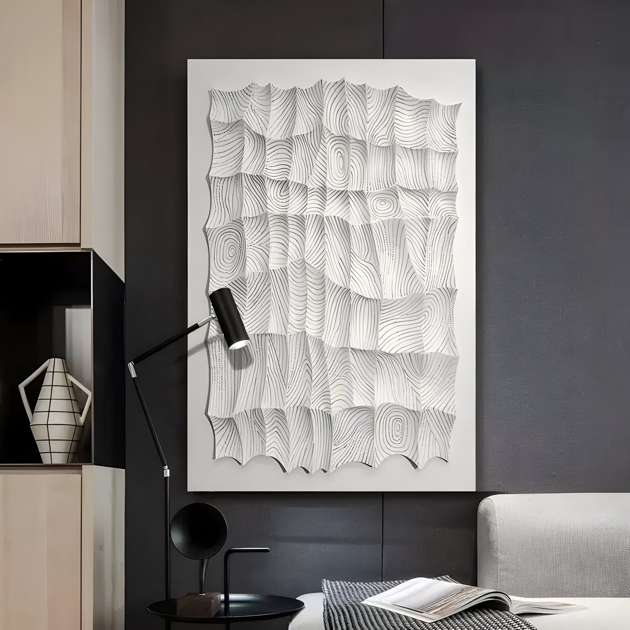 A modern room showcases the Novara White I Hand-Painted Wood Relief 3D Wall Art by Giant Sculptures, featuring black wood grain-like patterns. Nearby are a black desk lamp and chair, a geometric vase on a shelf, and part of a gray sofa.