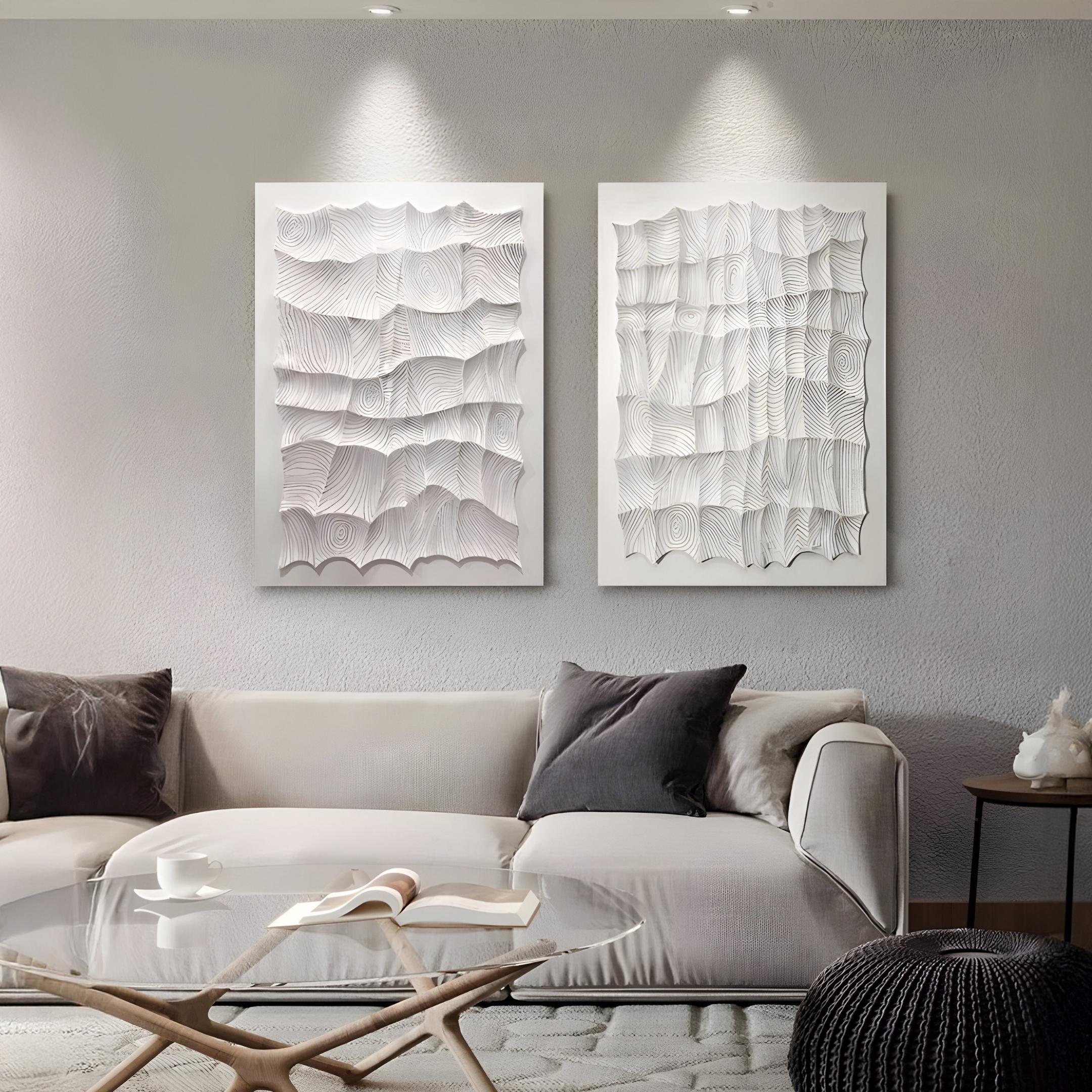A modern living room features a light gray sofa, wood and light throw pillows, a glass coffee table, and two Novara White II Hand-Painted Wood Relief 3D Wall Art pieces by Giant Sculptures above the sofa. Soft lighting highlights these works—ideal for minimal interiors.