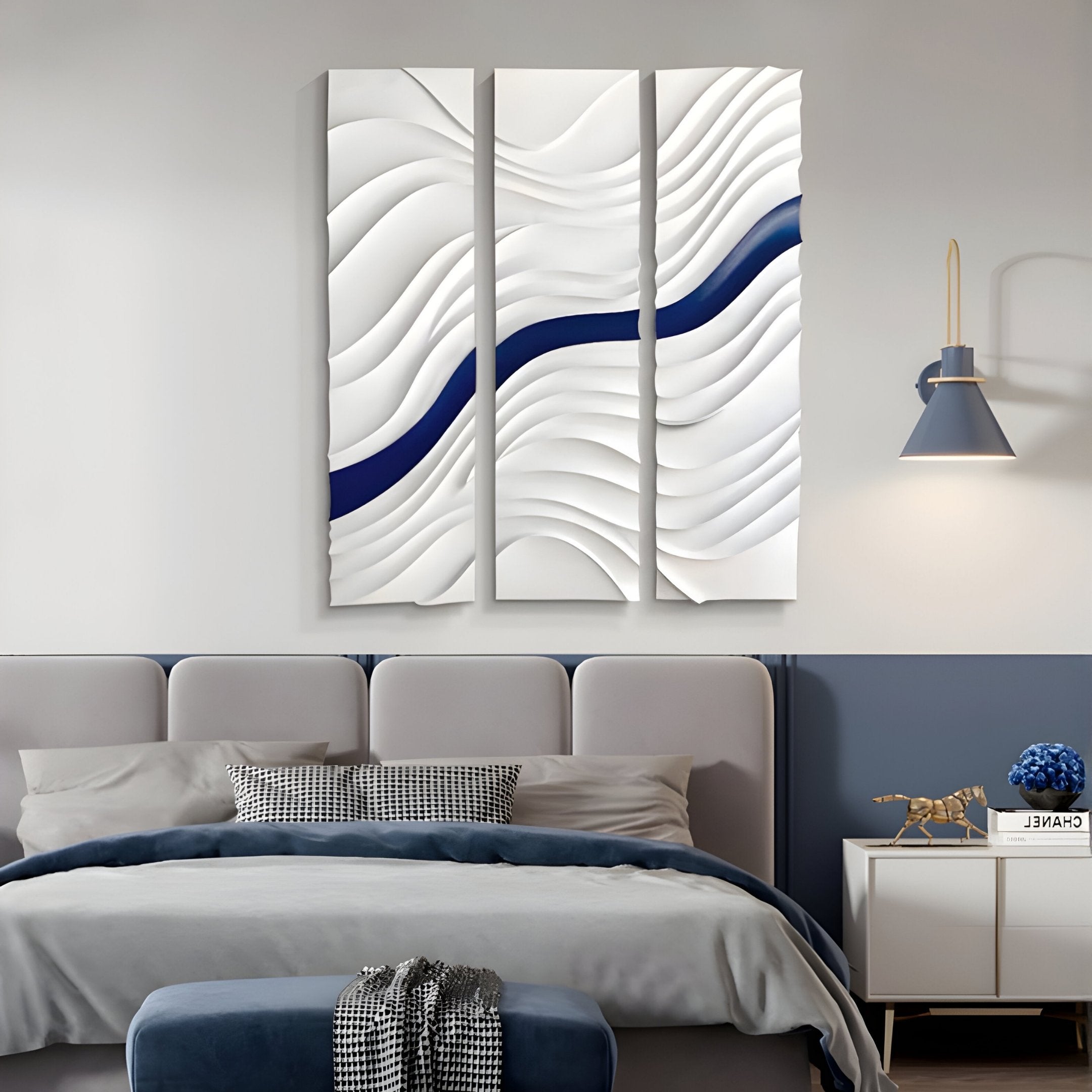 A modern bedroom features a gray bed with blue and white bedding, a blue ottoman, a bedside table with a blue vase, and the Evolva 3-Panel White & Blue Wood Relief 3D Wall Art by Giant Sculptures above the bed.
