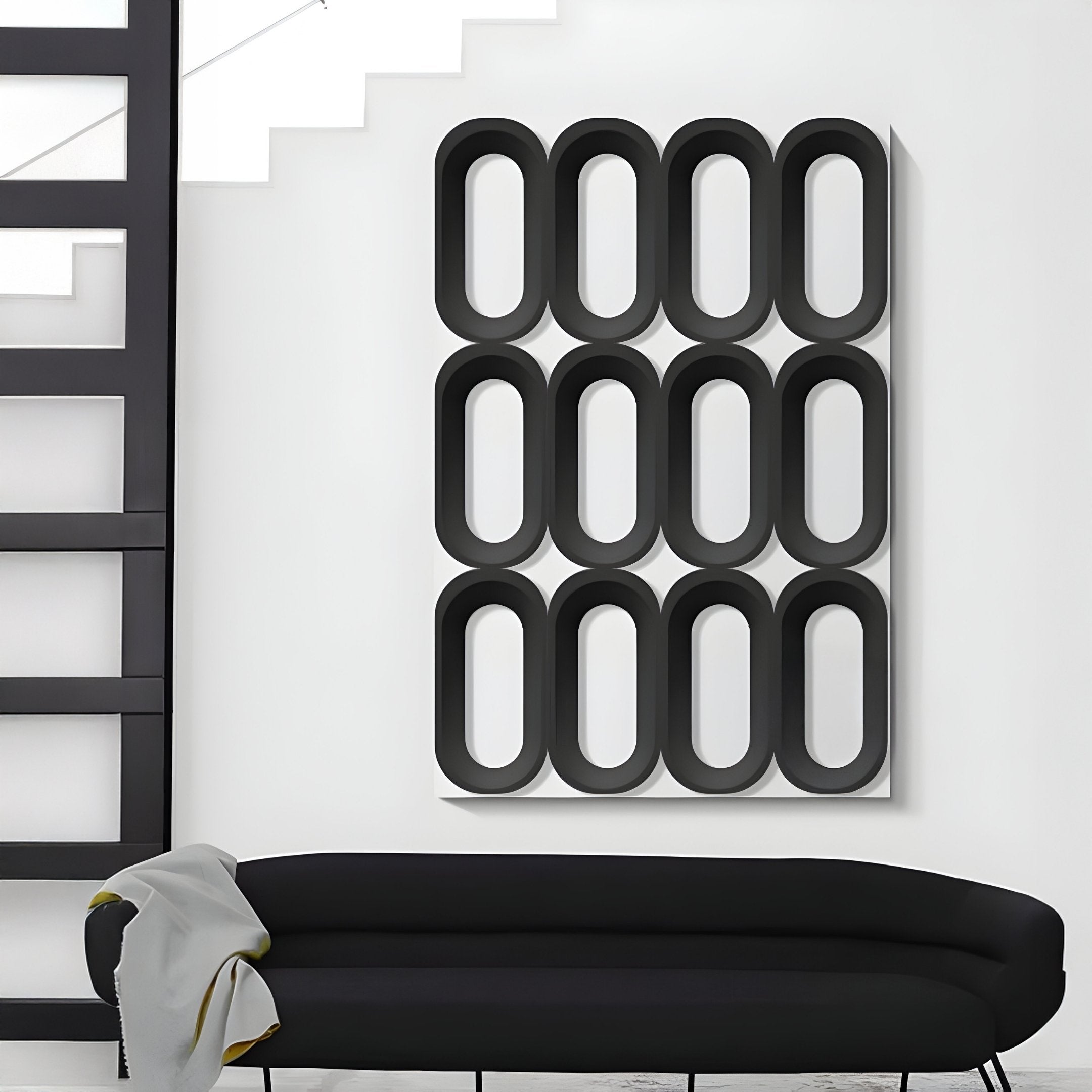 A modern living room with a black curved sofa and gray blanket, accented by Giant Sculptures Revela White & Black Geometric Wood Relief 3D Wall Art in a grid pattern. Stairs and a black frame appear on the left.