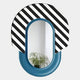The Arcus Blue & Black-White Striped Mirror 3D Wooden Wall Art by Giant Sculptures features an oval design with a bold striped top and solid blue base; a green leafy plant is reflected in this striking piece.