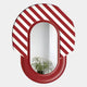 The Arcus Red & White Striped Mirror 3D Wooden Wall Art by Giant Sculptures features a bold oval frame with vivid diagonal stripes, reflecting a potted plant and crisp white walls in the background.