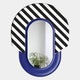 The Arcus Purple & Black-White Striped Mirror 3D Wooden Wall Art by Giant Sculptures features a graphic frame with bold stripes above smooth solid blue, reflecting a plant and white walls in the background.