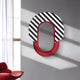 The Arcus Red & Black-White Striped Mirror 3D Wooden Wall Art by Giant Sculptures boasts a modern oval design, perfect for contemporary interiors. Mounted on a gray wall, it casts shadows near a window draped with white curtains and a partially visible red chair in the room.