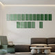 A modern living room features the Eximia Green Tones Multi-Panel Wood Relief 3D Wall Art by Giant Sculptures above a dark sofa. A minimalist coffee table with stacked books sits in front, while a uniquely shaped green chair complements the neutral-toned walls and floor.
