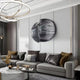 A modern living room features a spacious gray sectional with pillows and is highlighted by the Argentum Textured Wood Relief 3D Wall Art With Silver Metal Ball by Giant Sculptures on the gray wall. A round coffee table below a contemporary chandelier completes the neutral-toned setting.