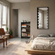A cozy, modern bedroom with a low wooden bed, beige bedding, large windows, indoor plants, and a bookshelf. The Giant Sculptures Lumiscale Black & White Modular Glass Wall Mirror adds contemporary style as natural light fills the space.