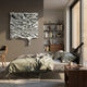 A modern bedroom features a neatly made bed, wooden nightstand, bookshelves, and large windows. Above the bed hangs the Lumineris Silver Melted Wave Metal 3D Wall Art by Giant Sculptures, adding striking abstract metal decor.