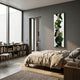 A modern bedroom with a large window, neatly made bed, low bookshelf of books, chair, and the Verdurelle Green Layered Flow Felt 3D Wall Art by Giant Sculptures adds abstract green flair to the gray wall. Natural light fills this minimalist space.