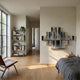A modern, minimalist bedroom with light wood floors, a gray bedspread, a wooden chair, tall bookshelf, potted plant, and Giant Sculptures' Auresmap Silver Cascade Multi-Panel Waterfall Wall Art as striking decor; sunlight streams in through large windows.