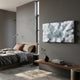A modern, minimalist bedroom features a gray bed, brown and beige pillows, a wooden bench with books and a vase, and Giant Sculptures’ Nivara Ice Blue Ripple Straw 3D Wall Art. Sunlight streams in through tall windows, warming the cozy interior.