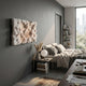 A modern bedroom with a gray accent wall, the Nivara Warm Beige Ripple Straw 3D Wall Art by Giant Sculptures, a neatly made bed with neutral bedding, a nightstand with books and lamp, and large windows overlooking city buildings and trees.