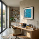 A modern home office with a wooden desk, laptop, books, and lamp. Large windows brighten the space. Shelves line the back wall, while Giant Sculptures' Trevasse Aqua Blue Textured Abstract Framed Wall Art hangs above the stylish chair.