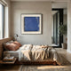 A cozy bedroom features a neatly made bed with earth-toned bedding, a bedside table holding books and a cup, wall-mounted lamp, large window, and Trevasse Deep Blue Textured Abstract Framed Wall Art by Giant Sculptures above the bed. A potted plant sits by the glass door.