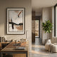 A modern home office features a wooden desk with a laptop, books, and mug. The Bravura I Stroke Abstract Framed Canvas Wall Art by Giant Sculptures is displayed on the wall, while a leafy plant sits by a cozy chair near large sunlit windows.