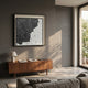 A modern living room with a gray wall, Stratonis Graphite Linework Textured Framed Wall Art by Giant Sculptures, wooden sideboard with books and lamp, beige sofa, and sunlight through glass doors opening to a green balcony.