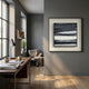 A modern home office features a wooden desk, books, coffee mug, lamp, and potted plant by a large window. An armchair with a blanket sits nearby. Stratonis Monochrome Layered Textured Framed Wall Art by Giant Sculptures decorates the gray wall.