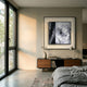Modern bedroom with large window, gray walls, and a bed with gray bedding. A wooden sideboard holds decor and books, while Stratonis Grey Terrain Textured Framed Wall Art by Giant Sculptures hangs above it, sunlight casting shadows in the minimalist space.