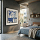 A modern bedroom with minimalist decor features the Stratonis Blue & Beige Grid Abstract Textured Framed Wall Art by Giant Sculptures, a wooden bed with gray bedding, bookshelves, and expansive windows for natural light.