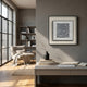 A modern, minimalist home office features a gray wall with the Stratonis Grey Linework Textured Framed Wall Art by Giant Sculptures, books, and a black vase on the desk. Sunlight streams in from large windows; a cozy chair and bookshelf complete the scene.