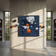 A modern gallery with tall windows showcases the Aeroluxe Midnight Blue Abstract 3D Wall Art With Metal Balls by Giant Sculptures. Orange, white, and blue 3D spheres emerge from the piece, adding energy among bookshelves along the walls.