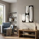 A cozy living room with a blue armchair, beige blanket, wooden coffee table, bookshelf full of books, and natural light from a window. The wall features the Curventa Black & White Curved Panel 3D Wall Art by Giant Sculptures.